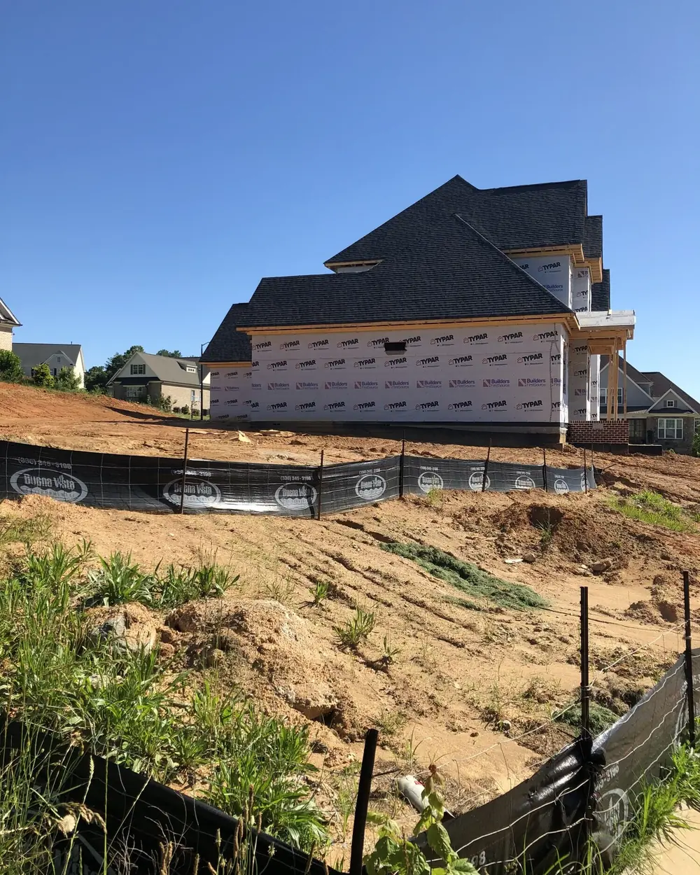 A two-storey home under construction on a graded hillside lot, dark roof on and sheathing up